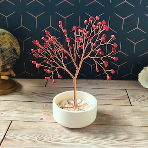 Op de afbeelding: Een decoratieve koperdraadboom met rode edelsteenbladeren in een witte keramische pot. De boom staat op een houten oppervlak, met een wereldbol en een patroonachtergrond zichtbaar.