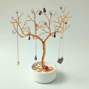 Puede incluir: Un soporte para joyas con forma de árbol de alambre de color cobre con una base de cerámica blanca. El árbol exhibe varios collares, pendientes y anillos. La base está llena de piedras blancas y una pequeña mariquita roja. El soporte para joyas está sobre un fondo blanco.