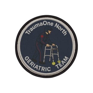 Peut inclure: Un écusson circulaire avec le texte "TraumaOne North GERIATRIC TEAM" en lettres blanches. L'écusson présente une illustration d'une personne utilisant un déambulateur. Le fond est bleu marine avec une bordure noire et blanche.