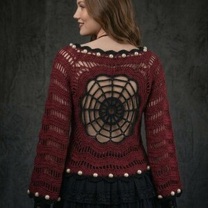 May include: A long-sleeved crochet sweater in deep burgundy featuring a prominent black spiderweb design on the back. The garment is accented with white pearl-like beads along the neckline and hem, and it is paired with a black skirt.