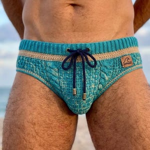 Pode incluir: Uma sunga de crochê turquesa com textura de tricô trançado, uma faixa horizontal bege na cintura e um cordão azul-marinho escuro com ponteiras metálicas. Um pequeno emblema de couro marrom com um ícone de onda está na lateral.