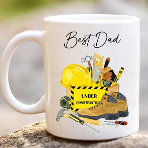 May include: White ceramic mug with a handle. The mug has a graphic of a yellow hard hat, tools, and work boots in a wooden box. The text "Best Dad" is printed in black cursive above the graphic. The text "Under Construction" is printed in black on a yellow warning sign.