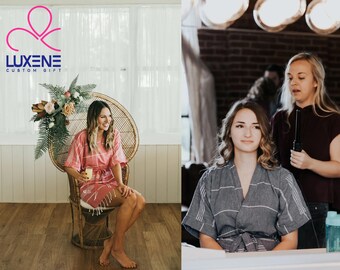 Può includere: Due donne in un ambiente di preparazione. Una donna indossa una vestaglia a righe rosa e siede su una sedia di vimini. L'altra donna indossa una vestaglia a righe grigie e si sta facendo acconciare i capelli. Il testo "LUXENE CUSTOM GIFT" è visibile nell'angolo in alto a sinistra dell'immagine.