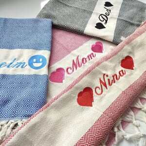 Op de afbeelding: Vier Turkse handdoeken met geborduurde ontwerpen. Een handdoek is blauw met een witte streep en een smiley, een andere is roze met een witte streep en het woord "Mam", een andere is wit met rode strepen en het woord "Nina" en de laatste is zwart-wit met het woord "Papa".