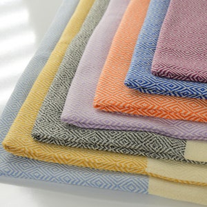 Puede incluir: Una pila de toallas turcas coloridas con un patrón de diamante. Las toallas son en tonos de morado, azul, naranja, lavanda, gris, amarillo y azul claro.