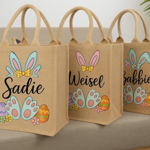 May include: Three burlap tote bags with bunny ears and paw prints. The bags are personalized with the names Sadie, Weisel, and Babbie. The bags are decorated with colorful Easter eggs and flowers.