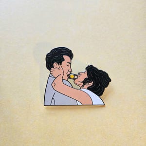 以下が含まれることがあります： ロマンチックな抱擁をするカップルを描いたピンバッジ。一人が黄色い物をもう一人に食べさせている。人物は黒い線で縁取られ、男性はグレーのシャツ、女性は白いトップスを着ている。背景は薄い黄色。