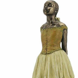 May include: A bronze sculpture of a young ballerina wearing a yellow bodice and a white tutu. The ballerina is standing on a small base and is looking up.