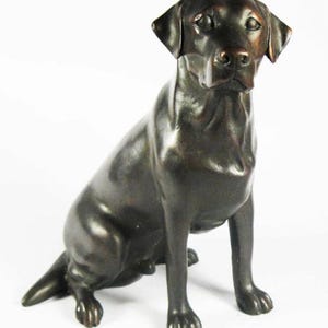 May include: A bronze sculpture of a black Labrador Retriever sitting with its head tilted slightly to the side. The dog has a realistic appearance with detailed features.