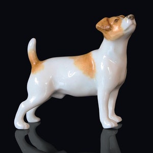 May include: A white porcelain figurine of a Jack Russell terrier dog with brown markings. The dog is standing on all four legs with its head tilted up.