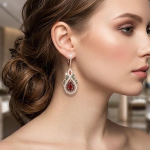 May include: A rose gold drop earring featuring a central pear-shaped red gemstone surrounded by a halo of small clear crystals, accented with green and red stones in an ornate filigree frame, suspended from a crystal-encrusted lever back hook.