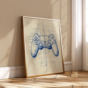 May include: A framed print featuring a blue blueprint-style illustration of a video game controller. The controller is centered on a beige background, with a gold frame. The artwork is leaning against a wall.
