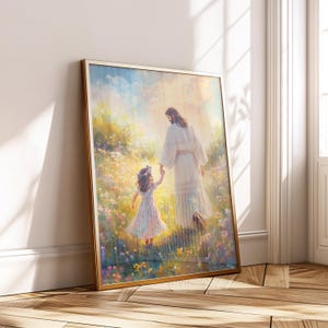 May include: A framed art print depicts a figure in white robes holding hands with a young girl in a field of flowers. The artwork has a gold frame and is leaning against a white wall. The scene is bathed in soft light, creating a serene and uplifting atmosphere.