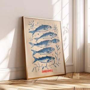 May include: Framed art print featuring six blue sardines with detailed scales and fins, arranged vertically. The print includes botanical elements and the word "SARDINES" in red. The artwork is displayed in a gold frame.