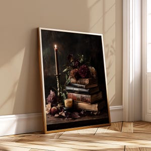 May include: A framed art print featuring a still life composition. A lit candle sits in a brass candlestick next to a stack of old books and burgundy flowers. The artwork has a dark, moody aesthetic.