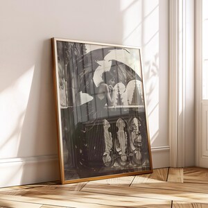 Puede incluir: Impresión enmarcada en blanco y negro que representa a una mujer en un balcón con un paraguas. La obra de arte tiene una estética vintage, con detalles arquitectónicos y un estilo gótico. El marco es dorado.