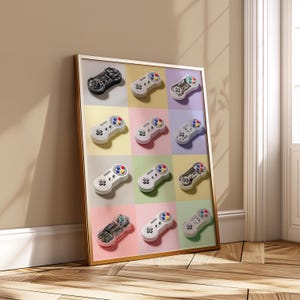 May include: A framed print featuring a grid of colorful squares, each displaying a different retro video game controller. The controllers vary in color and design, with a gold frame. The background is a neutral beige.