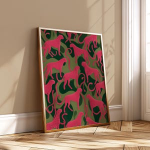 May include: A framed art print featuring a repeating pattern of pink cheetahs on a green and dark green camouflage background. The artwork is displayed in a gold frame and leans against a wall.
