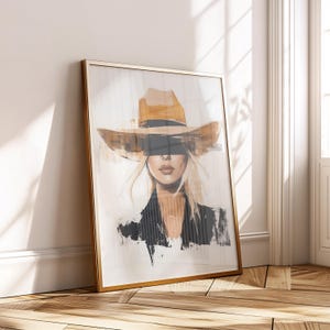 May include: Framed art print featuring a woman wearing a tan cowboy hat. The artwork has a watercolor style with a gold frame. The woman has blonde hair and is wearing a black blazer. The art is leaning against a white wall.