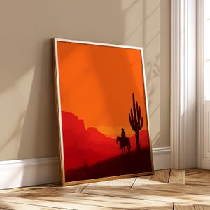 Puede incluir: Impresión artística enmarcada que representa a un vaquero a caballo, silueteado contra una vibrante puesta de sol naranja y roja. Un gran cactus se alza a la derecha. La obra de arte está en un marco dorado, apoyada contra una pared.