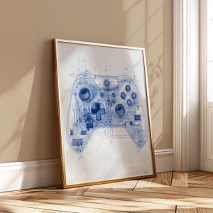 May include: A framed print featuring a detailed blueprint of a video game controller. The controller design is rendered in blue lines against a white background, with a gold frame. The artwork is leaning against a wall.