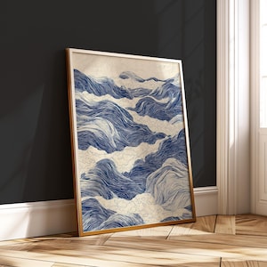 May include: A framed art print featuring an abstract design of blue and white waves. The artwork is in a gold frame and leans against a dark wall. The print's design is reminiscent of a flowing ocean or river.