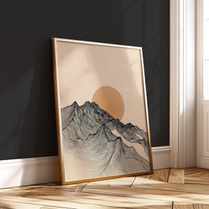 May include: Framed art print featuring a minimalist mountain landscape with a rising sun. The artwork showcases black line art against a beige background, encased in a gold-colored frame. The print leans against a dark wall, adding a touch of modern decor.