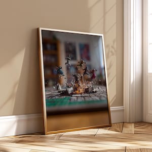 Puede incluir: Una impresión enmarcada que presenta figuras en miniatura en el aire sobre una mesa, con un efecto de explosión. Las figuras son de varios colores y el marco tiene un acabado dorado. El fondo está borroso, lo que sugiere un entorno de habitación.