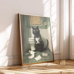 Op de afbeelding: Een ingelijste kunstdruk toont een zwarte kat in een badkamer, omringd door verspreid toiletpapier. De kat, met grote ogen, houdt een prop papier vast bij een toilet. Het kunstwerk heeft een gouden lijst en leunt tegen een witte muur.