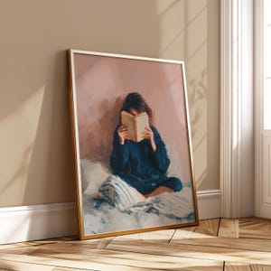 May include: A framed art print showing a person reading a book. The person is wearing a dark blue sweater and sitting on a bed with a striped pillow. The artwork has a soft, painterly style and is presented in a gold frame.