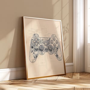 Può includere: Una stampa incorniciata con un progetto dettagliato di un controller per videogiochi. Il design del controller è in bianco e nero, mostrando i componenti interni e la disposizione dei pulsanti. La stampa ha uno sfondo beige e una cornice dorata, appoggiata a un muro.