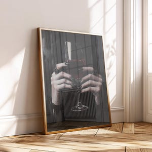 Könnte beinhalten: Gerahmter Kunstdruck mit einem Weinglas, gefüllt mit roter Flüssigkeit, gehalten von Händen mit Ringen und dunklem Nagellack. Das Kunstwerk hat einen goldenen Rahmen und eine dunkle, stimmungsvolle Ästhetik.