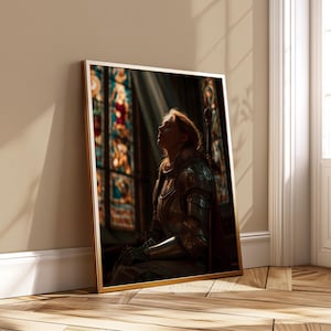 May include: A framed print featuring a person in medieval armor, illuminated by light from a stained-glass window. The artwork has a gold frame and is leaning against a wall. The person is looking up, with a sword resting behind them.