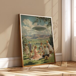 May include: Framed print of a retro picnic scene with a family on a grassy hill. A silver UFO hovers above. The artwork has a vintage aesthetic with a gold frame and a landscape background, reminiscent of the 1950s.