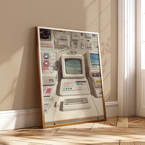Puede incluir: Una impresión enmarcada de una sala de control de computadoras retro. La obra de arte presenta un monitor de computadora blanco, un teclado y varios paneles de control en una habitación de color claro. La impresión está en un marco dorado y se apoya contra una pared.