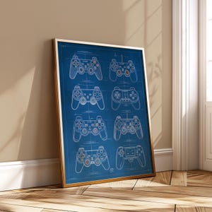 May include: A framed print showing blueprint-style illustrations of several video game controllers. The controllers are white on a dark blue background, with a gold frame. The print is leaning against a wall and floor.