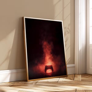 May include: Framed art print depicting a video game controller against a red, smoky backdrop. The controller is centrally positioned, within a gold frame. The artwork is leaning against a beige wall and wooden floor.