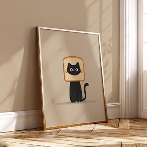 Puede incluir: Un cuadro enmarcado con un gato negro con una rebanada de pan tostado en la cabeza. El gato tiene ojos negros y una cola curvada. El fondo es de color beige neutro y el marco es dorado.