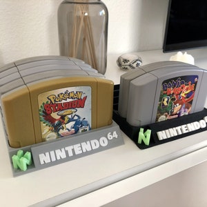 Op de afbeelding: Twee Nintendo 64-spelcartridgehouders, een grijs en een zwart, met het Nintendo 64-logo gedrukt op de voorkant. De grijze houder heeft een stapel van zes spelcartridges, waaronder "Pokémon Stadium". De zwarte houder heeft twee spelcartridges, waaronder "Banjo-Kazooie".