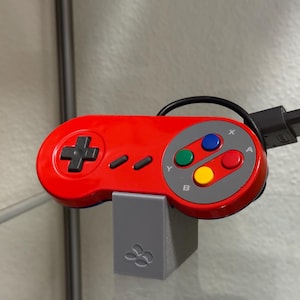 Könnte beinhalten: Ein roter Videospiel-Controller mit grauem Tastenlayout und schwarzem Kabel und Stecker. Der Controller hat ein Steuerkreuz, Tasten und einen grauen Ständer mit Logo.