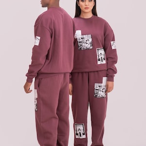 May include: Burgundy sweatshirt and sweatpants set. The sweatshirt and sweatpants have patches with black and white graphics. The set is worn with white clogs. The set is made of a soft material.