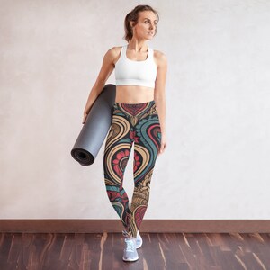 May include: A woman wearing a white sports bra and patterned leggings. The leggings feature a colorful design with red, blue, and gold floral and abstract shapes. She is holding a rolled-up gray yoga mat.
