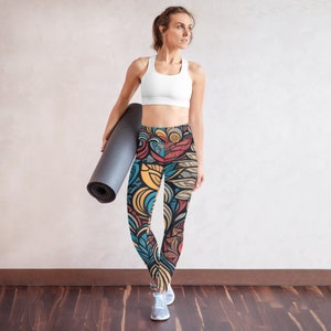 May include: A woman wearing a white sports bra and patterned leggings. The leggings feature a colorful floral design with orange, blue, and yellow accents. She is holding a gray yoga mat and wearing gray sneakers.