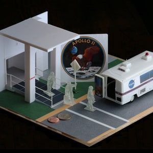 Op de afbeelding: Een miniatuur diorama dat de Apollo 11-missie uitbeeldt. Het toont een witte NASA-bus, een lanceerstructuur en astronautenfiguren. Het Apollo 11-embleem is zichtbaar. De scène is geplaatst op een grijze en groene basis.