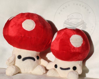 May include: Two plush red and white mushroom toys with white faces and black eyes. The larger mushroom has a sad expression, while the smaller mushroom has a happy expression.
