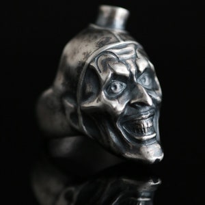 Puede incluir: Un anillo de plata con una escultura detallada de la cara de un bufón con una amplia sonrisa y un sombrero puntiagudo. El anillo está sobre un fondo negro.