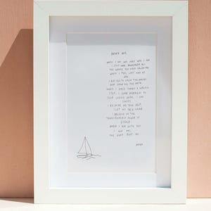 May include: A white framed print with handwritten text and a small sailboat illustration. The text begins with "Father God," and continues with a prayer. The frame is white, and the background is a pale peach colour.