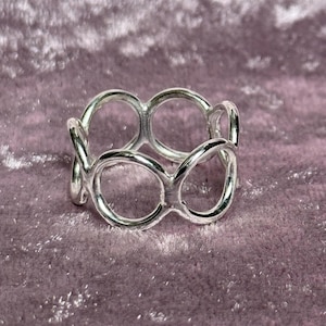 Puede incluir: Un anillo plateado con un diseño único que presenta formas circulares interconectadas. El anillo está hecho de un metal brillante y reflectante y se encuentra sobre una superficie púrpura texturizada. El diseño del anillo es abierto y aireado.
