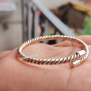 May include: A twisted metal bracelet with a silver and gold finish. The bracelet has a spiral design and a clasp closure. The bracelet is shown on a person's hand. The text "StoreForRings" is visible.