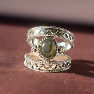 Pode incluir: Um anel de prata com uma gema verde engastada em um bezel decorativo. O anel é de dupla banda com um design ondulado.